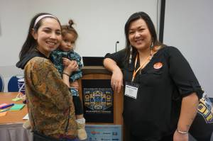 Annauk Olin, holding her daugher Tulġuna Taas Olin, and Rochelle Adams pose on March 20, 2024, after giving a presentation on language at the Alaska Just Transition Summit in Juneau. The two, who work together at the Alaska Public Interest Research Groups Language Access program, hope to compile an Indigenous environmental glossary. (Photo by Yereth Rosen/Alaska Beacon)