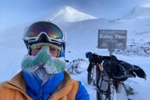 Peter Delamere poses in front of a sign in Rainy Pass in the Alaska Range during the 2024 Iditarod Trail Invitational race. On his face is a nose-hat invented by Fairbanks athlete Shalane Frost. (Photo by Peter Delamere)