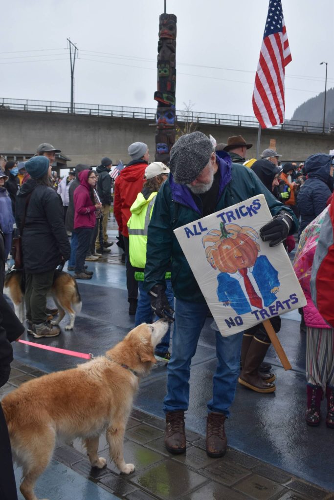 A dog sniffs the hand of a protester holding a sign that reads, All Tricks, No Treats, depicting President Donald Trump as a pumpkin. (Mari Kanagy/Juneau Empire)